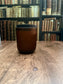 Brown cylindrical candle  jar on a wooden surface with a bookshelf in the background