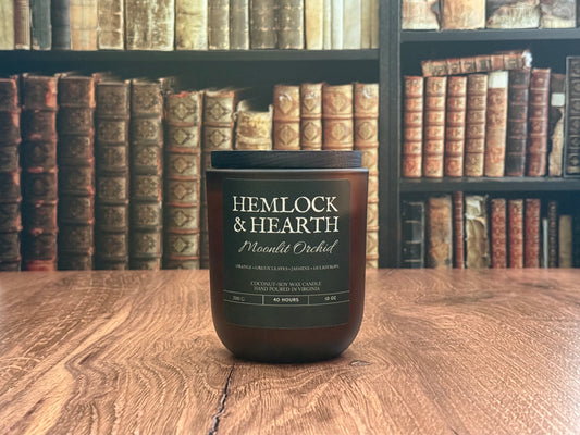 Candle labeled 'Hemlock & Hearth' on a wooden surface with bookshelves in the background